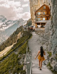 a woman is walking up the stairs to a house on top of a mountain with mountains in the background