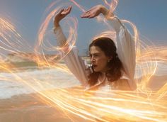 a woman standing in the water with her arms spread out and fire spinning around her