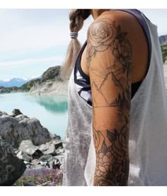 a woman with a flower tattoo on her arm standing next to a lake in the mountains