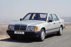 a white mercedes benz is parked on the side of the road in front of some hills