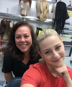 two women sitting at a table in front of wigs and hairbrushes on shelves