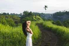 a woman in a white dress standing on a dirt road near tall grass and palm trees