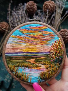 someone is holding up a small embroidery art piece that looks like a sunset over a lake