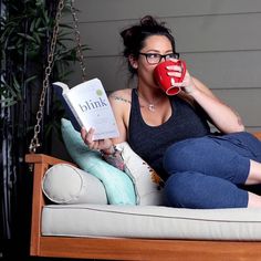 a woman sitting on a porch swing holding a book and drinking from a red mug