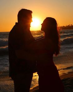 a man and woman standing on top of a beach next to the ocean at sunset