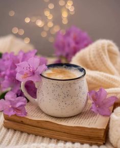 a cup of coffee and some purple flowers on a wooden book with a white knitted blanket in the background