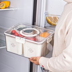 a woman holding a plastic container in front of an open refrigerator with fruit and vegetables inside