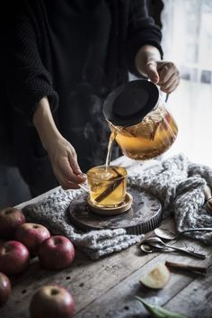 a person pouring tea into a cup on top of a wooden table next to apples