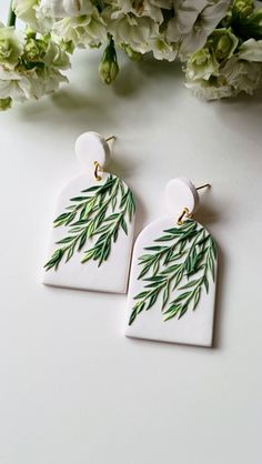 two white ceramic earrings with green leaves on them, sitting next to some flowers and greenery