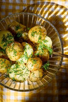 a glass bowl filled with potatoes covered in dill sprinkles on a yellow and white checkered tablecloth