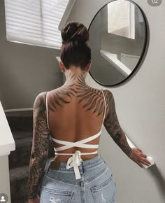 a woman with tattoos on her back standing in front of a mirror and looking at herself