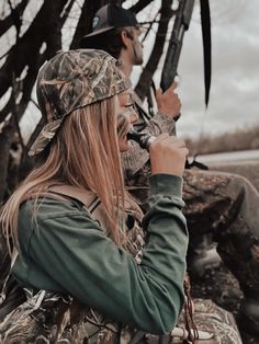 a man and woman sitting next to each other in front of trees with hunting gear