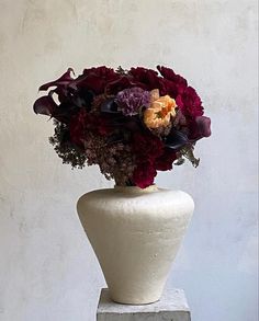 a white vase filled with lots of flowers on top of a cement block in front of a wall