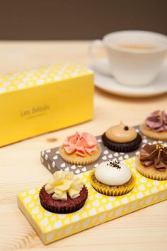 there are many different cupcakes on the table next to a yellow box with white polka dots