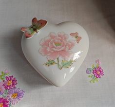 a white heart shaped box with pink flowers on it and butterflies around the edges, sitting on a tablecloth