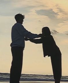 two people are standing on the beach and holding hands with each other as the sun sets