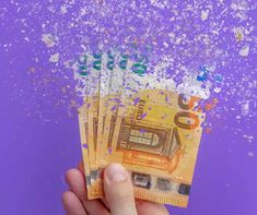 a hand holding five bills in front of a purple background with white speckles