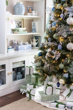 a christmas tree with presents under it in front of a white hutch filled with blue and green ornaments