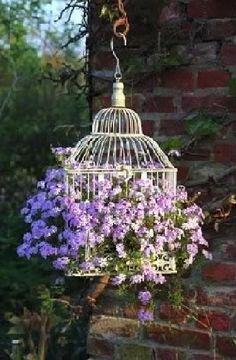 a white birdcage with purple flowers hanging from it's side next to a brick wall