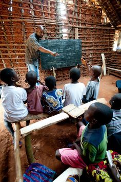 a man standing in front of a group of children sitting on the ground next to a chalkboard