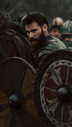 a man with a beard standing next to another man in armor and holding a shield