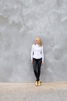 a woman standing in front of a gray wall wearing black pants and a white shirt