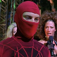 a man in a red spider suit is talking into a microphone while two women stand behind him