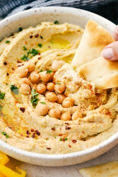 a hand dipping hummus into a white bowl with pita chips on the side