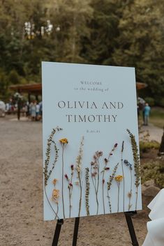 a sign with flowers on it in the middle of a dirt road next to trees