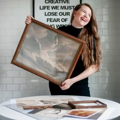 a woman holding up a framed photo on top of a table with other pictures around it