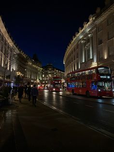 two red double decker buses driving down a street next to tall buildings at night time