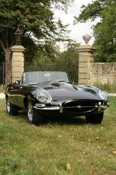 an old black sports car parked in front of a stone wall and gated entrance