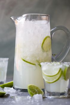 a pitcher filled with ice and limes next to two glasses full of lemonade