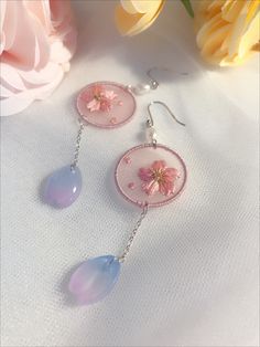 two pink earrings with flowers on them are sitting next to a flowery vase and white cloth