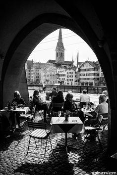 black and white photograph of people sitting at tables under an arch in a city area