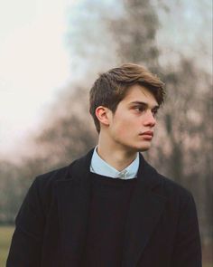 a young man standing in front of trees wearing a black coat and blue shirt with his hands in his pockets