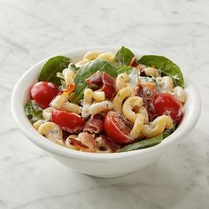 a white bowl filled with pasta salad on top of a marble counter topped with tomatoes and spinach