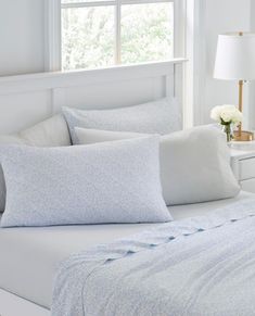 a bed with white sheets and pillows in front of a window next to a night stand