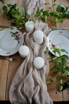 the table is set with four white candles and two plates on it, along with greenery