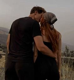 a man and woman standing next to each other in front of a fence with mountains behind them