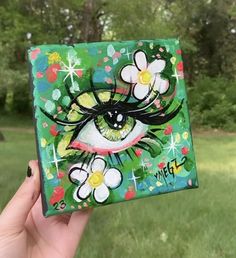 a hand holding up a small square piece of art with flowers and an eye painted on it