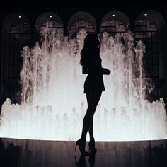 a woman standing in front of a fountain with water shooting up from it's sides