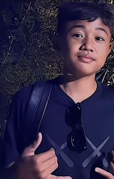 a young boy wearing a black shirt and tie standing in front of some bushes with one hand on his hip