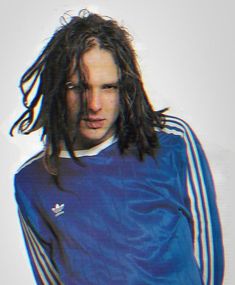 a man with dreadlocks standing in front of a white wall wearing a blue adidas shirt