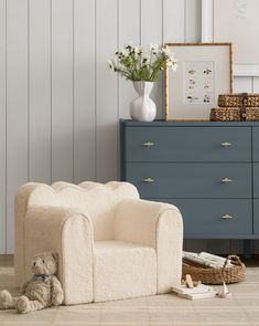 a teddy bear sitting on the floor next to a chair and dresser with flowers in it