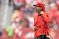 a man wearing a red hoodie and black pants on the sidelines during a football game