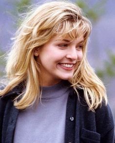 a woman with long blonde hair smiles at the camera while wearing a gray sweater and black jacket