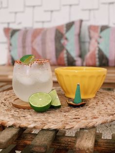 two margaritas sitting on top of a wooden table next to a yellow bowl and lime