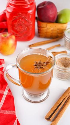 a cup of apple cider next to cinnamon sticks, apples and other ingredients on a white surface