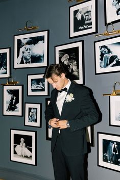 a man in a tuxedo looking down at his watch while standing next to a wall with pictures on it